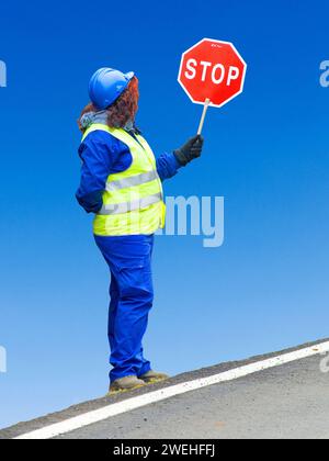 Woman in yellow signal waistcoat and blue safety helmet stands at the side of the road holding up a stop sign Stock Photo