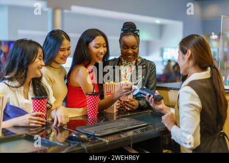 Diverse group of female friends, representing different nationalities, joyfully uses credit cards to pay for movie tickets at the theater Stock Photo