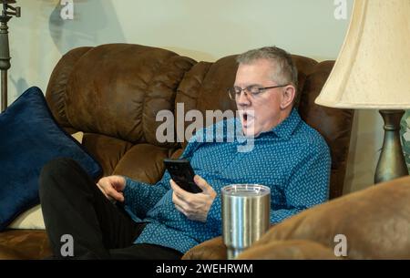 60 year old adult Caucasian man relaxing on sofa and staring at something on his phone with shock or surprise. Stock Photo
