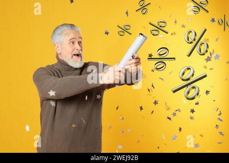 Discount offer. Happy senior man blowing up party popper on golden background. Confetti and percent signs in air Stock Photo