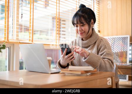A positive, beautiful Asian woman in a cosy sweater is responding to messages or reading texts on her phone while working remotely at a coffee shop. p Stock Photo