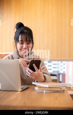 A positive, beautiful Asian woman in a cosy sweater is responding to messages or reading texts on her phone while working remotely at a coffee shop. p Stock Photo