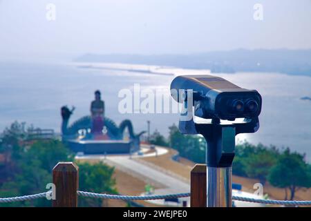 Samcheok City, South Korea - December 28, 2023: Observatory binoculars in the foreground, with a blurred silhouette of the Lady Suro sculpture and the Stock Photo