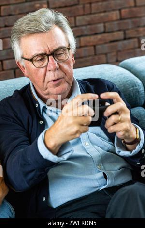 The image depicts an absorbed senior man engaged with his smartphone, possibly immersed in a game or app. His focused gaze and the intent way he interacts with the device suggests concentration and perhaps enjoyment of modern digital entertainment. The casual yet smart attire of a button-up shirt under a sweater indicates a relaxed setting, possibly at home, where comfort meets a touch of formality. This photo represents the growing trend of seniors adopting and enjoying new technology for leisure. Senior Man Concentrated on Smartphone Gaming. High quality photo Stock Photo