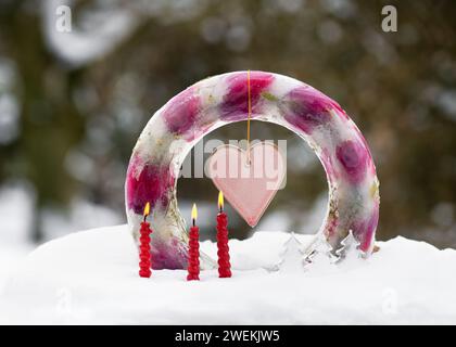 Handmade frozen wreath in shape of ring cake made of ice, pink tulip flowers with pink heart and burning red candles in snow. Stock Photo