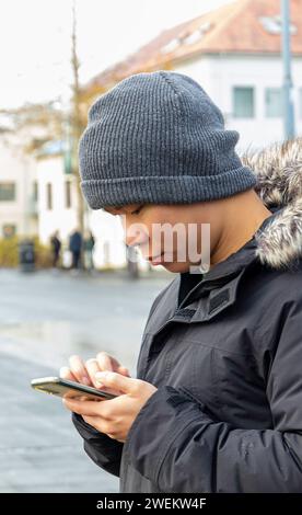 Side view of smiling young male in warm clothes looking at screen of mobile phone while standing on city road and checking for text messages against b Stock Photo