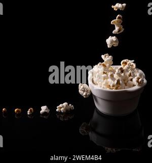 Popcorn falls into a white ceramic bowl and on the shiny black background with reflection Stock Photo