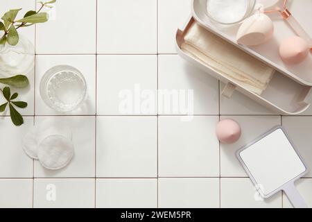 Makeup sponge, massage roller in plastic box with drawer, mirror and cotton pads and green leaves on white tile floor. Blank background with lifestyle Stock Photo