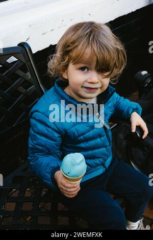 Little boy in blue coat eats blue ice cream on city bench Stock Photo