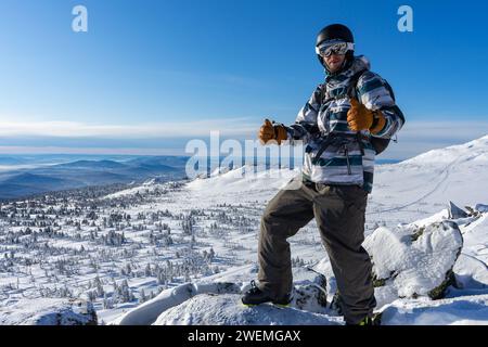 Happy smiling man in skiing winter equipment shows thumbs up, recommendation right choice. Warm jacket, pants, brown gloves, ski goggles, boots, sport Stock Photo