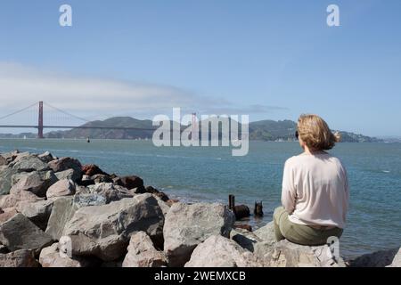 Young woman viewing the iconic Golden Gate Bridge spanning the Golden Gate in San Francisco, California from a viewing point. Stock Photo