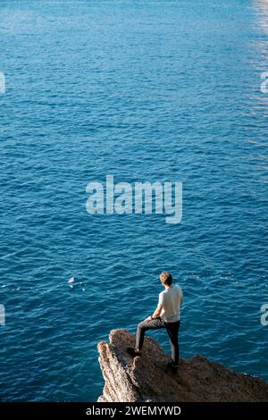 Young man stands on a stone ledge above the sea and looks into the distance. Back view Stock Photo