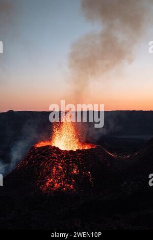 A photograph capturing the eruption of the Piton de la Fournaise volcano as it spews lava against a backdrop of a setting sun. Stock Photo