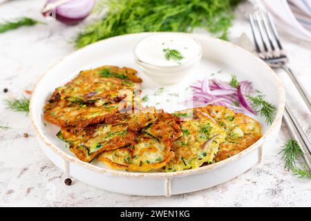 Zucchini fritters with red onions, garlic and herbs. Vegetarian zucchini pancakes and sour cream on white table. Stock Photo