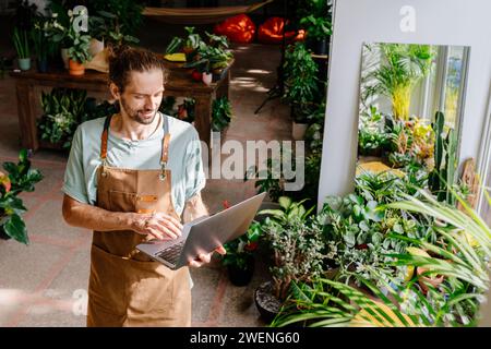 Caucasian guy wearing apron working with laptop in a home indoor. Man working with laptop or computer in cozy room with greenery plants. Stock Photo