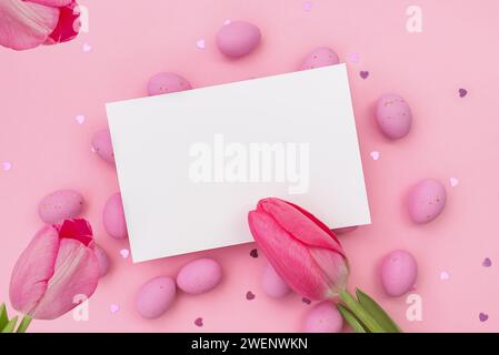 Easter postcard. Invitation mockup or blank with White card for greeting or invitation lies on Easter chocolate pink eggs. Three Red tulips. Shimmering glowing hearts. Happy holidays. Copy space Stock Photo