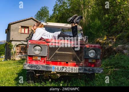 A woman looks thoughtfully at the sky over the hood of an old red off-road vehicle, sideways Stock Photo