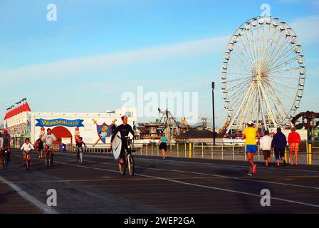 An adult man rides his bicycle while carrying his surfboard in the boardwalk in front of a Ferris Wheel, in Ocean Stock Photo