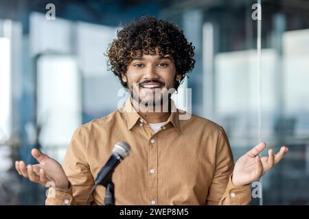 Handsome arab businessman sitting in stylish office and speaking in front of microphone. Positive male employer giving supportive speech for coworkers on corporative event and gesturing with hands. Stock Photo