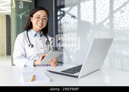 Joyful asian intern woman dressed in medical coat sitting at desktop with laptop and holding digital tablet in hands. Smiling female nurse using modern gadgets for productive work in hospital office. Stock Photo