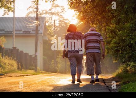 An elderly couple is walking along the street holding hands. Walk of old people at sunset on a summer evening. Stock Photo