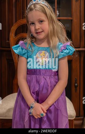 Four year old blonde Caucasian girl standing in front of a chair while dressed in a Disney costume, Tinkerbell,  jewelry & tiara while role playing. USA Stock Photo