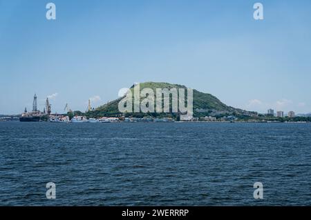 Distant view of Morro da Penha hill in Niteroi's Ponta Dareia district as saw from Guanabara bay blue waters under summer afternoon clear blue sky. Stock Photo