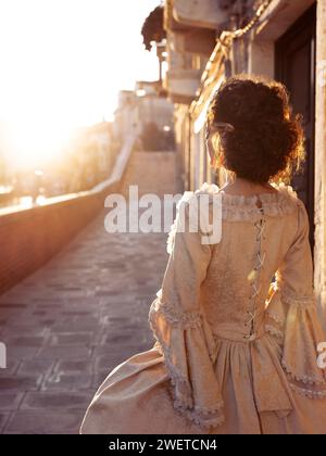 Artistic outdoor portrait of a beautiful lady wearing a Venetian carnival mask and an elegant vintage Italian masquerade ball dress walking along a su Stock Photo