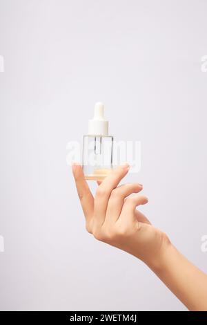 Glass serum bottle with dropper cap in woman's hand. Labelless clear glass bottle containing colorless essence on light background. Mockup for cosmeti Stock Photo