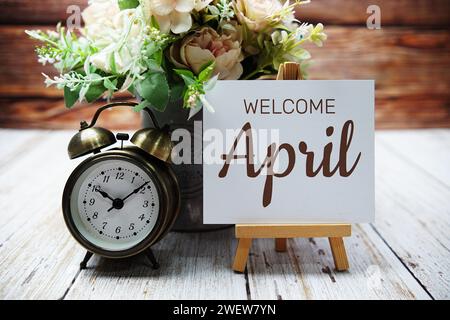 Welcome April text message written on paper card with wooden easel and alarm clock with flower in metal vase decoration Stock Photo