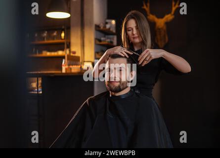 Young long haired female barber gently makes undercut hairstyle using scissors and comb bearded happy man in Salon Chair. Modern black-style barber sh Stock Photo