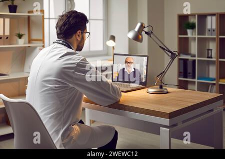Doctor in white coat, sitting at his workplace in the office during online meeting using webcam video call on laptop computer, communicates with Stock Photo