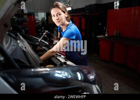 Young woman works under the open hood of a car, she has a special tool in her hands Stock Photo