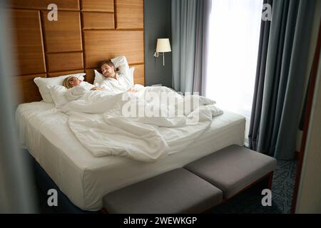 Blonde with her boyfriend are talking in a cozy hotel room, they are sitting on a large bed Stock Photo