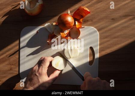 Male hands slicing onion with a knife on a kitchen table, on white cutting board, with sunlight from window, top view Stock Photo