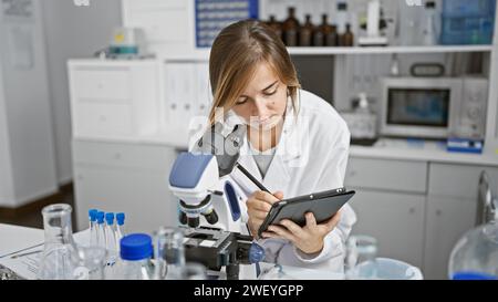 Attractive young blonde scientist, close-up portrait of her concentrated work on analysis and research in the lab, using microscope and writing on the Stock Photo