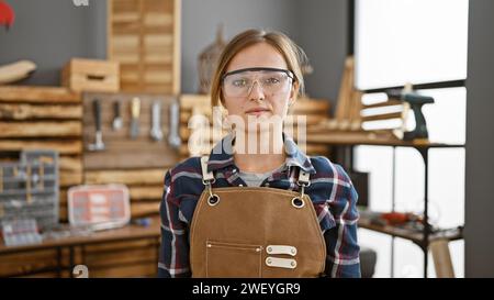 Attractive young blonde female carpenter in security glasses, a portrait of serious professionalism inside her bustling carpentry workshop. Stock Photo