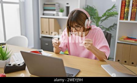 Captured in concentration, a young, beautiful hispanic woman worker, immersed in her business work inside the office, working hard with laptop and hea Stock Photo
