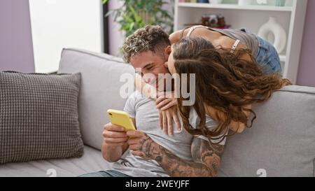 Affectionate woman embraces smiling tattooed man using smartphone on a cozy living room couch Stock Photo