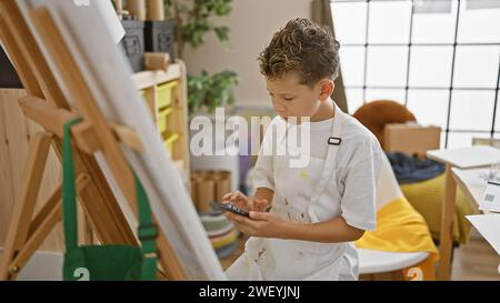 Adorable blond boy artist earnestly looking at smartphone, learning to draw in cozy art studio class Stock Photo