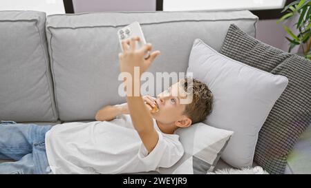 Adorable blond boy seriously engrossed, lying on sofa at home, using smartphone technology to watch video, eating a delicious croissant snack Stock Photo