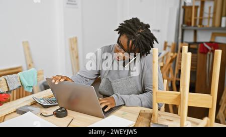 Young black woman with dreadlocks multitasking in a carpentry workshop, using a laptop while talking on the phone. Stock Photo