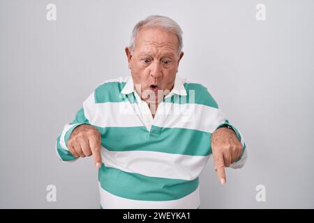 Senior man with grey hair standing over white background pointing down with fingers showing advertisement, surprised face and open mouth Stock Photo