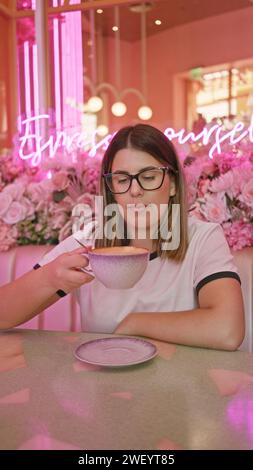 Adult hispanic woman enjoying coffee in a pink-themed cafe with neon lights and floral decorations. Stock Photo