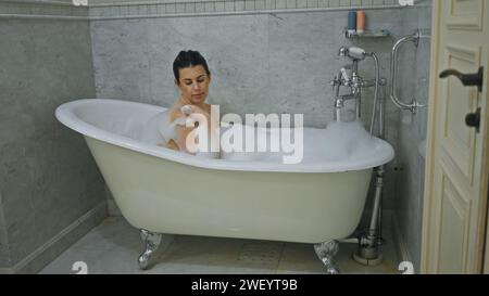 A relaxed woman enjoys a bubble bath in an elegant clawfoot bathtub within a marble-tiled bathroom Stock Photo