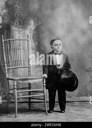 Nineteen year old little person man poses by a chair in a studio portrait, ca. 1895. Stock Photo