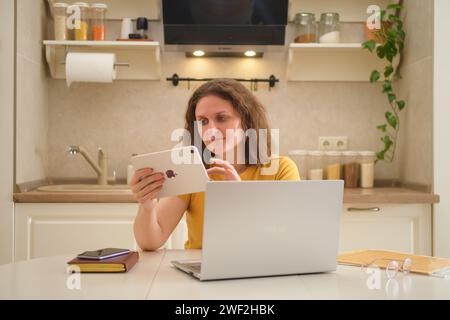 A woman with a laptop ASUS holds a digital tablet Apple iPad from a headache while sitting at a table in a home kitchen - Moscow, Russia, January 16, Stock Photo