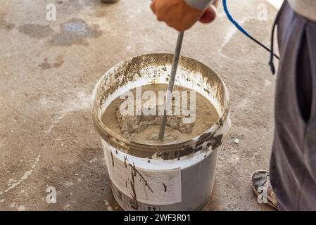 The worker mixes the solution in a bucket using a mixer. Mixing plaster solution in a bucket using electric drill. Stock Photo