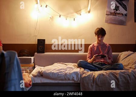 Wide angle portrait of teen boy playing mobile games sitting on bed with legs crossed in real room Stock Photo