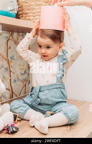 Little baby girl sitting on the table putting paper bucket on her head. Stock Photo